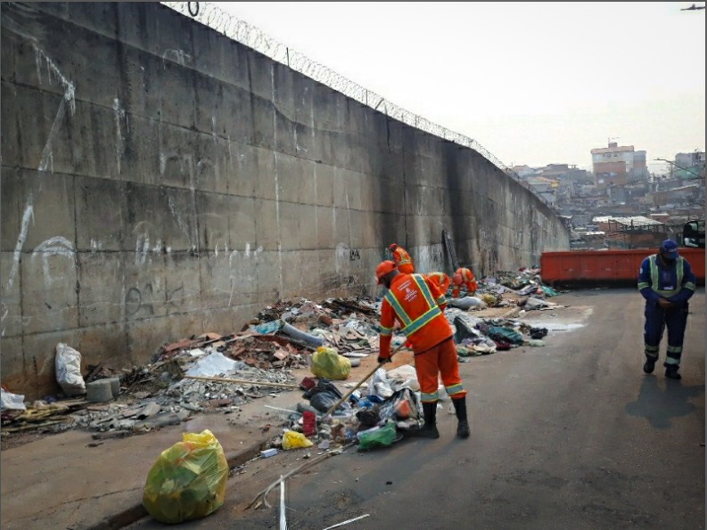 Em uma rua próxima a um grande muro com arame farpado, agentes de limpeza urbana realizam a remoção de lixo e entulho descartados irregularmente. Vestindo uniformes laranja com faixas refletivas, um trabalhador varre os resíduos, enquanto outros estão ao fundo coletando materiais. Uma grande caçamba laranja está posicionada para receber o descarte correto. No lado direito da imagem, um outro profissional, usando uniforme azul, acompanha a ação. Ao fundo, é possível ver uma comunidade com diversas casas em uma área elevada.